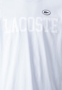 Balta medvilnė marškinėliai su dideliu ryškiu "LACOSTE" užrašu baltomis raidėmis ir mažuoju juodu siuvinėtu krokodilu baltoje fone.