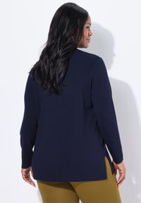 Frau mit langen schwarzen Haaren, die einen dunkelblauen Pullover und senfgelbe Hosen trägt, seitlich vor einem weißen Hintergrund.