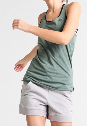 Kobieta w zielonym topie Adidas i szarych szortach stoi z jedną ręką zgiętą w swobodnej pozie na jasnym, jednolitym tle.