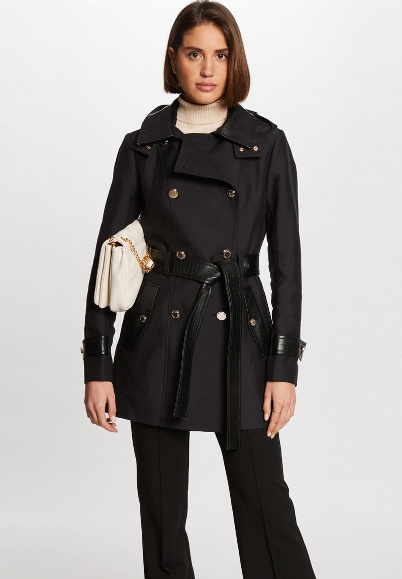 Svart dobbeltradet trenchcoat med belte i midjen, teksturerte svarte skinn detaljer og gullfargede knapper; sammen med en hvit håndveske.