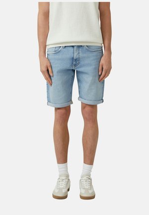 Person trägt hellblaue, hochgekrempelte Jeansshorts, weißes strukturiertes Top, weiße Socken und beige Sneaker und steht mit entspannten Händen.