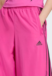 Pink athletic shorts with black side stripes, elastic waistband, and a side pocket. Features the Adidas logo in black on the lower leg.