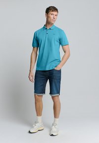 Turquoise poloshirt met korte mouwen, zwart logo, gecombineerd met opgerolde denimshorts en witte sneakers met grijze accenten.