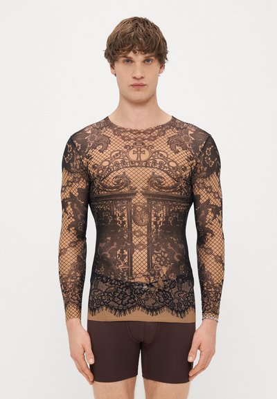 Langsleeve top in doorschijnend zwart gaas met ingewikkeld kant en houtpatroon, voorzien van een bruine onderlaag. Gecombineerd met bruine shorts.
