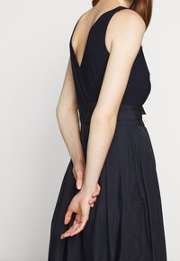 Kvinna iförd en ärmlös svart klänning med djup V-rygg, stående med händerna sammanfogade bakom ryggen mot en enfärgad bakgrund.