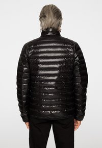 Svart puffjacka med en blank, vadderad textur och horisontella ränder. Hög krage och långa ärmar, matchad med svarta byxor.