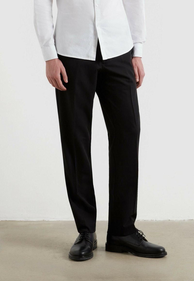 Czarne spodnie garniturowe o prostym kroju. Wykonane z gładkiego materiału, z delikatną fakturą. Połączone z czarnymi skórzanymi butami.