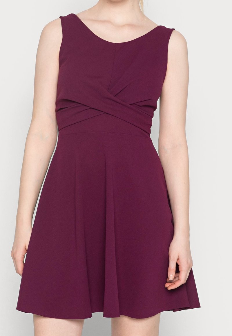 Robe sans manches de couleur prune avec un détail en tissu croisé à la taille et une jupe évasée, portée par une personne sur un fond uni.