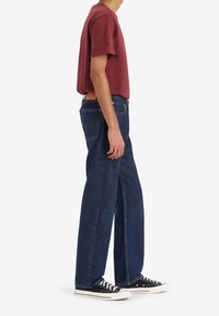 Bordeauxfärgad bomullströja i kombination med mörkblå vida jeans och svarta låga sneakers med kontrasterande vita sulor.