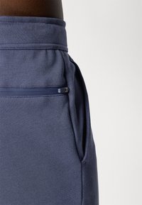 Zbliżenie na kieszeń granatowych spodni dresowych z poziomym zamkiem nad bocznym szwem.