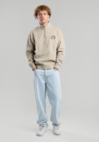 Beige pullover with a half-zip collar and black logo on chest, paired with light blue loose-fit jeans and white sneakers. Smooth texture.