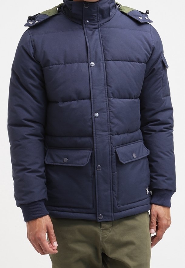 Dunkelblaue gesteppte Jacke mit Druckknopfleiste, zwei Klappentaschen, gerippten Bündchen und olivgrünen Schulterakzenten, getragen zu olivgrünen Hosen.