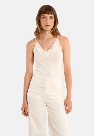 Donna con capelli castano chiaro che indossa una canotta bianca senza maniche con scollo a V e dettagli floreali, abbinata a pantaloni bianchi a vita alta, su uno sfondo neutro.