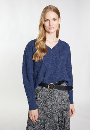 Dunkelblaues Strickpullover mit Rautenmuster, V-Ausschnitt, kombiniert mit einem gemusterten Rock und einem schwarzen Gürtel mit einer goldenen Schnalle.