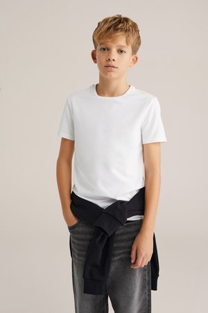 T-shirt blanc à manches courtes porté avec un pull sombre noué à la taille sur un jean noir délavé. Design simple, texture lisse, coupe décontractée.
