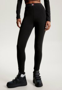 Černé elastické legíny s vysokým pasem a hladkou texturou. Obsahují nášivku s logem na pasu a jsou kombinovány s černými vysokými teniskami.