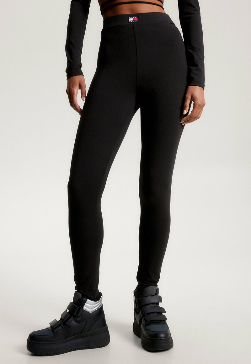 Černé elastické legíny s vysokým pasem a hladkou texturou. Obsahují nášivku s logem na pasu a jsou kombinovány s černými vysokými teniskami.