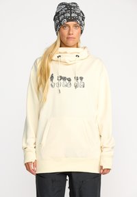 Sweatshirt à capuche crème avec cordon de serrage, comportant une poche avant et un texte graphique noir. Porté avec un bonnet noir à motifs.