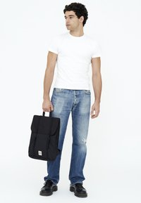 Svart ryggsäck med en vikbar klaff, två framremmar och en logopatch, hålls av en person i en vit t-shirt och slitna blå jeans.