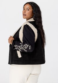 Zwarte vest met crème-kleurige nepbontafwerking en decoratieve borduursels op de mouwen. Model draagt crème-kleurige broek en heeft lang, golfachtig haar.