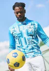 Maglietta sportiva azzurra chiara con collo a zip, caratterizzata da motivi geometrici e dettagli con il logo. Giocatore in pantaloni bianchi con un pallone da calcio giallo nelle vicinanze.