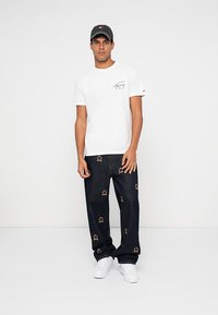 Hvit t-skjorte med logo, kombinert med mørkeblå jeans med krone-motiver. Brukt med hvite joggesko og en grå caps.