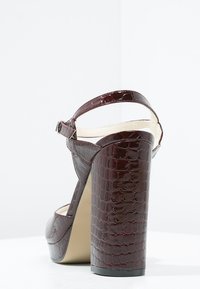 Glossy burgundy high-heeled sandal with a crocodile pattern, open back, ankle strap, and a chunky block heel. Sole is beige.