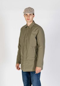 Giacca verde oliva con bottoni, collo a camicia, due tasche laterali e taglio dritto. Il modello indossa un berretto a quadri e jeans.