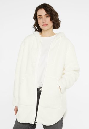 Young woman with short dark hair wearing a white fluffy hooded jacket over a white shirt and dark pants against a plain background.