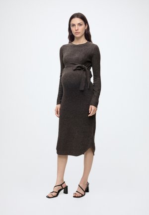 Femme enceinte portant une robe midi en maille côtelée marron foncé avec un nœud sur le côté, debout pieds nus sur un fond clair uni.