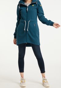 Felpa lunga color teal con cappuccio, dotata di chiusura con zip, due tasche frontali e lacci. Accentuata da un logo giallo sul petto.