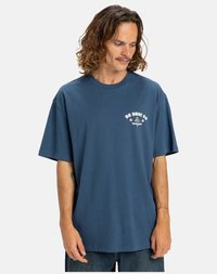 Man med axellångt lockigt hår, iklädd en lös blå DC Shoe Co t-shirt och jeans, ser nedåt och åt sidan.