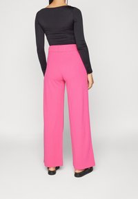 Pantalones de pierna ancha rosas con cintura elástica, combinados con una blusa negra de manga larga, mostrados desde atrás sobre un fondo neutro.