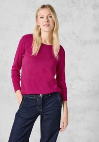 Maglione fucsia a maniche lunghe in tessuto morbido, abbinato a pantaloni in denim scuro con una vestibilità classica e dettagli di cucitura gialla.