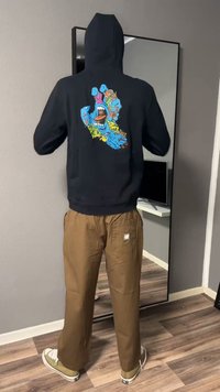 Persona che indossa una felpa nera con cappuccio con una grafica colorata di una mano blu con la bocca sulla schiena, pantaloni marroni e scarpe da ginnastica, in piedi al chiuso.