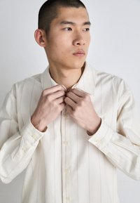 Camicia a maniche lunghe di colore crema, con righe verticali sottili, colletto a punta e dettagli con bottoni. Tessuto morbido.