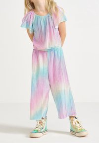 Colorful, pleated jumpsuit with short flutter sleeves in pastel pink, blue, and lavender. Paired with shiny high-top sneakers.