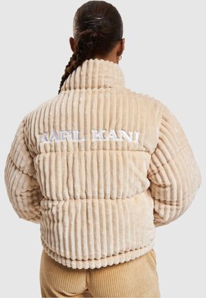 Mujer con el cabello trenzado que lleva una chaqueta de felpa beige con canalé con "KARL KANI" bordado en la espalda.