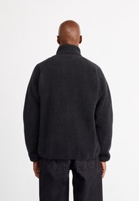 Black fleece jacket with a high collar, relaxed fit, and textured surface. Minimal design with no visible fastenings or accents.