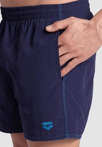Bañador navy hecho de un tejido ligero con cinturilla elástica, con costuras en azul brillante y un logo de "arena" en el dobladillo.