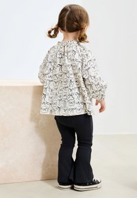 Long-sleeve top in cream with black bow print, paired with dark flared pants and black lace-up sneakers; textured fabric, relaxed fit.