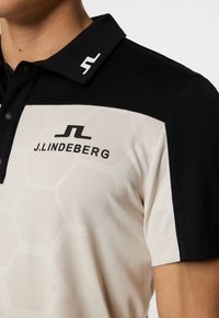 Kortärmad polo med svart krage, beige kropp med hexagonmönster och broderad J. Lindeberg-logga på bröstet.