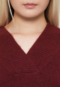 Zbliżenie dolnej części twarzy i szyi kobiety, która ma na sobie prążkowany, bordowy sweter z dekoltem w serek, z prostymi, blond włosami widocznymi.