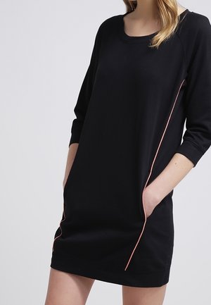 Femme portant une robe noire à manches longues avec des détails de passepoil rose, les mains dans les poches, sur un fond clair.