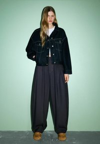 Schwarze Jeansjacke mit Fronttaschen und silbernen Knöpfen, kombiniert mit weiten dunklen Hosen und braunen Schuhen vor einem grünen Hintergrund.
