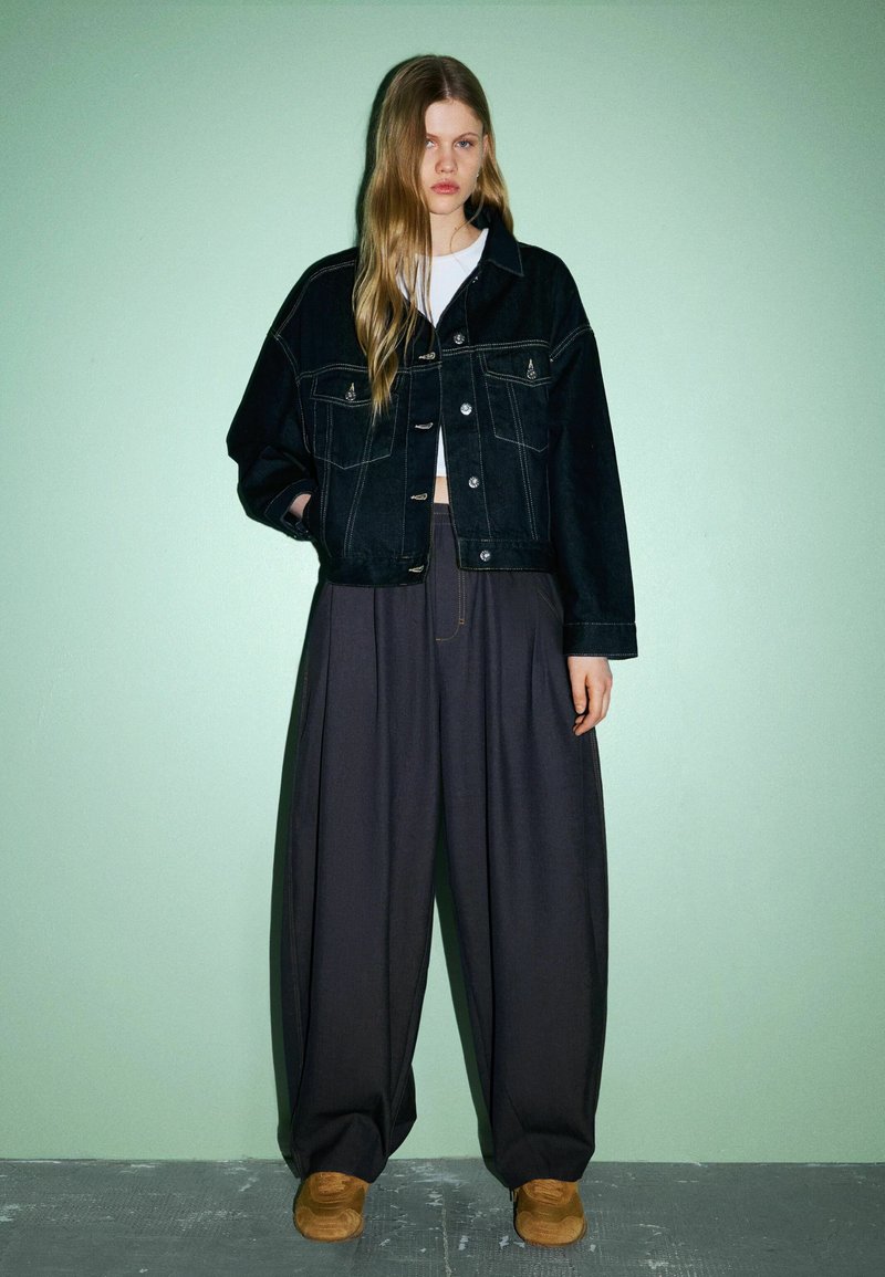 Schwarze Jeansjacke mit Fronttaschen und silbernen Knöpfen, kombiniert mit weiten dunklen Hosen und braunen Schuhen vor einem grünen Hintergrund.