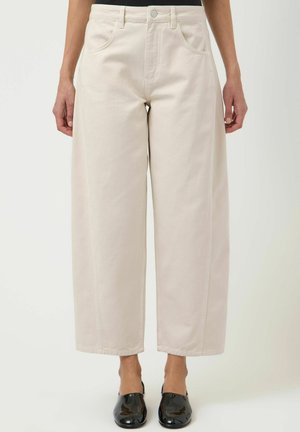Femme portant un pantalon taille haute, jambes larges, couleur crème, avec poches avant et des chaussures noires brillantes sans lacets, debout devant un fond uni.