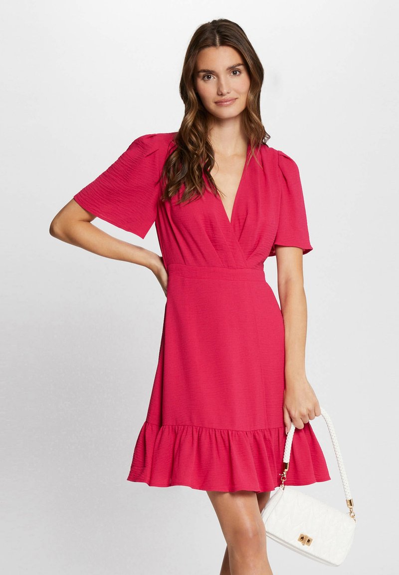 Korte roze jurk met een V-hals en puffmouwen; heeft een getailleerde taille en een gerimpelde zoom. Gecombineerd met een witte handtas met een gevlochten schouderband.