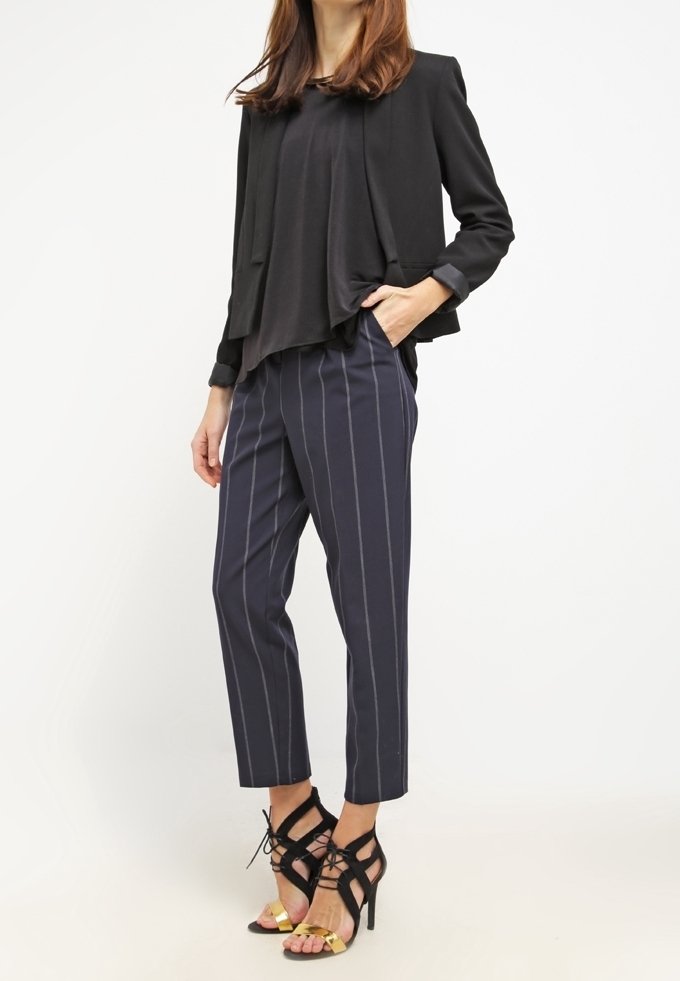 Femme portant un blazer noir et un top, un pantalon bleu marine à fines rayures et des sandales noires à talons hauts avec des accents dorés, debout sur fond blanc.