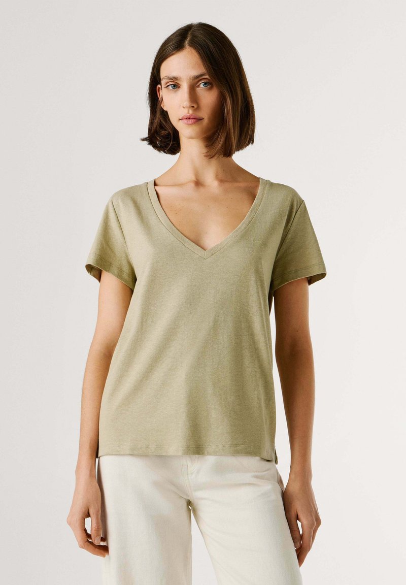 Femme aux cheveux bruns mi-longs portant un T-shirt vert olive clair à col en V et un pantalon blanc sur un fond uni.